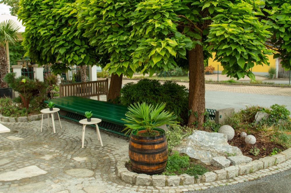 Schattiger Vorplatz mit grüner Bank, Holzfass und Kopfsteinpflaster