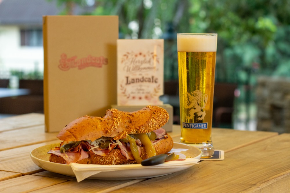 Belegtes Sesamweckerl mit einem Glas Bier neben der Willkommenskarte des Landcafe